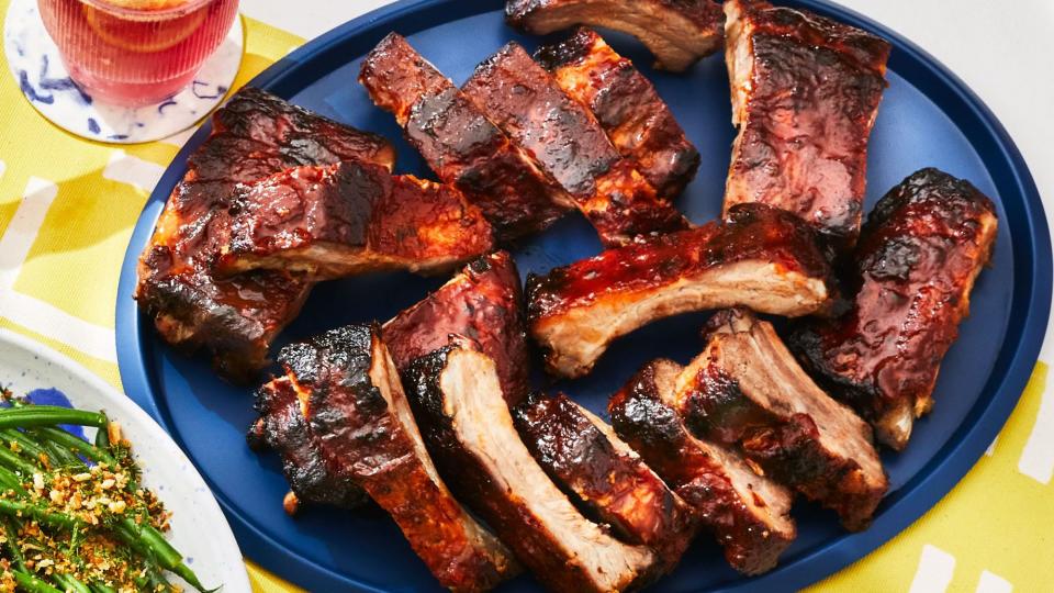 Platter of BBQ ribs set on a yellow print tablecloth with green beans and a red beverage.