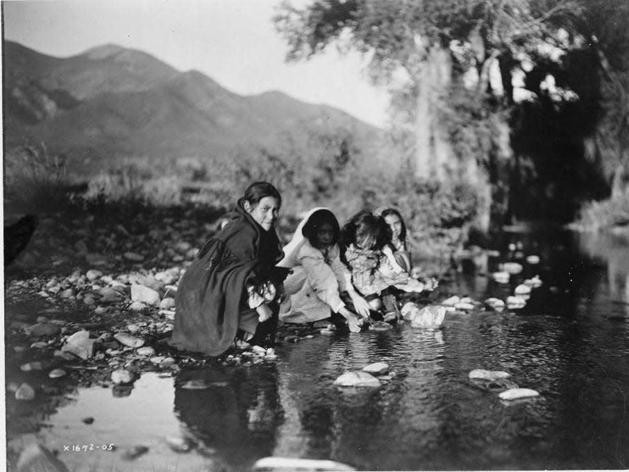 Four Taos children squat on rocks at edge of stream, mountains in background.