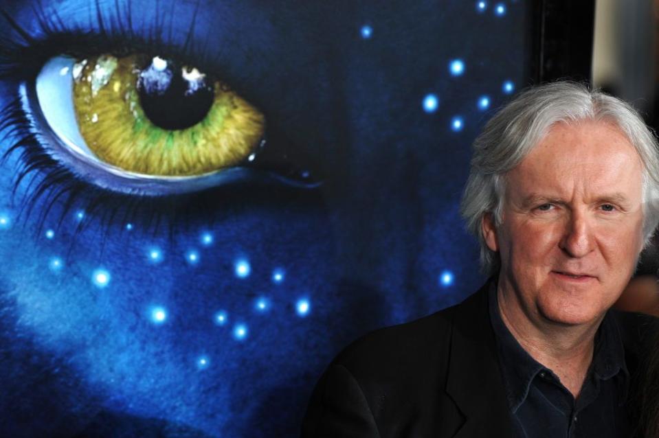 james cameron standing in front of a large avatar movie poster showing a closeup of a blue skinned alien with a yellow eye