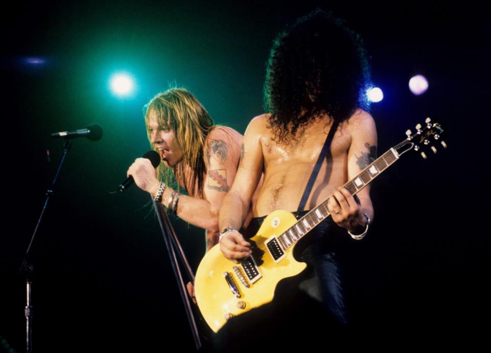 A man with long blond hair sings next to a man with black curly hair covering his face while he plays the guitar.