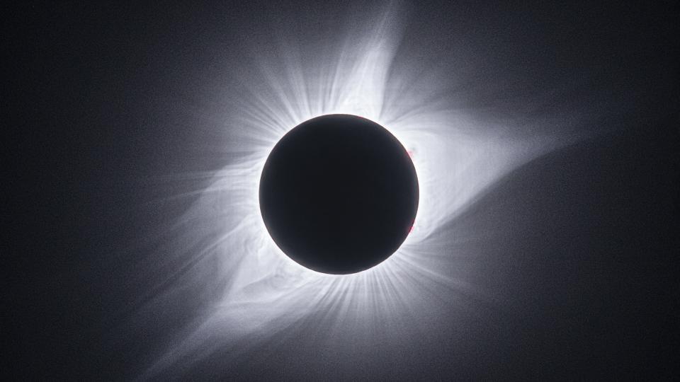total solar eclipse with dark circle in the center and white streamers and eruptions surrounding it.