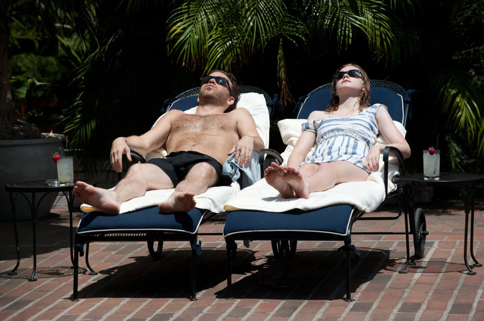 Elle Fanning lays poolside in a chaise chair. A man lays on his own chair next to her. They both wear dark sunglasses.