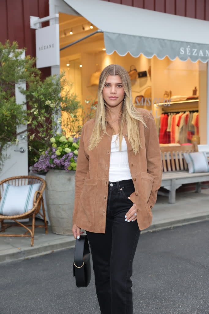 sofia richie poses for a photo while standing in front of a store, she wears a brown jacket over a white top and black jeans
