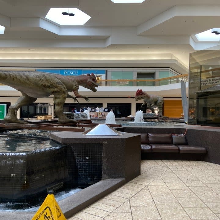 An empty mall with large animatronic dinosaurs in the center near a fountain