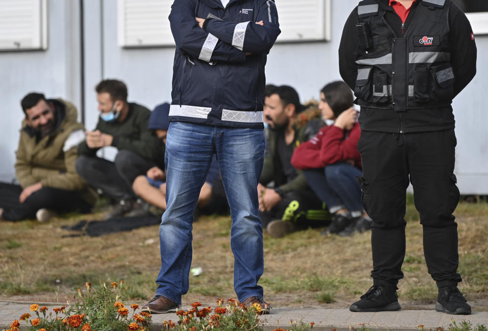 FILE-In this Oct. 6, 2021 taken photo security guards stand in front of migrants at the Central Initial Reception Facility for Asylum Seekers (ZABH) in the state of Brandenburg in Eisenhuettenstadt, Germany. German authorities say that the number of migrants arriving in the country via Poland and Belarus has gone up quickly during recent months. (Patrick Pleul/dpa via AP)