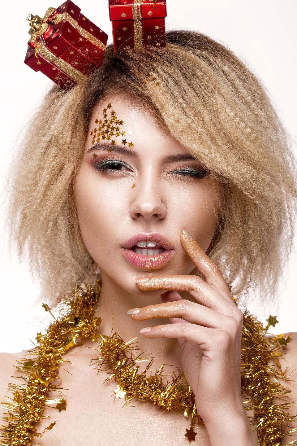 woman with crimped blonde hair winking at camera with gold stars above one eyebrow and two red and gold wrapped christmas presents on an angle on her head with gold tinsel around her neck