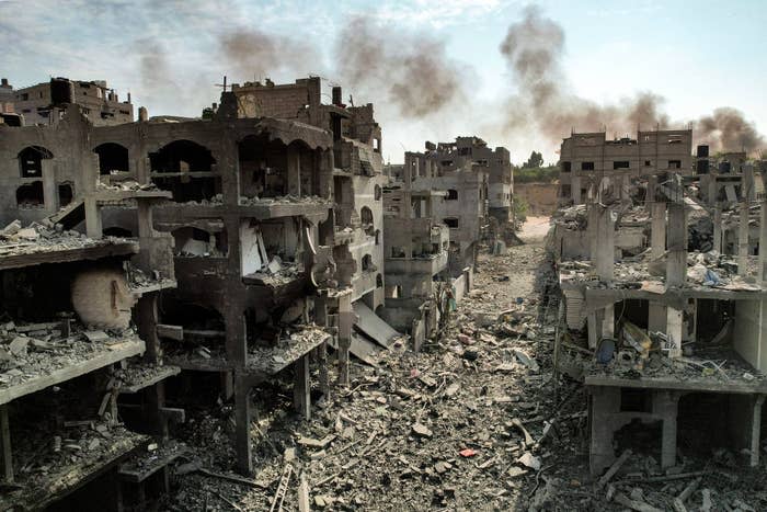 Ruined buildings and debris after likely conflict, with smoke rising in the background