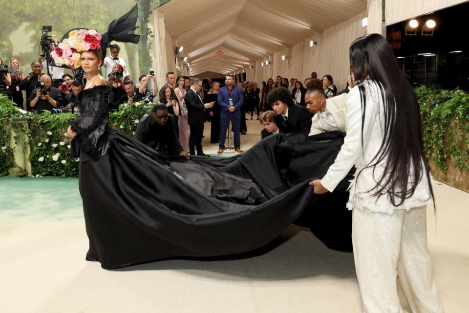 Zendaya in a long gown with a floral headpiece on red carpet, assisted with her train by Law