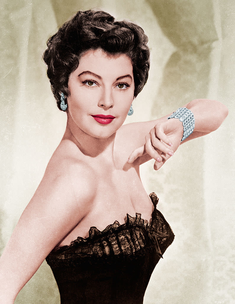 Ava Gardner is wearing a strapless, lace-trimmed dress and a sparkling bracelet, posing with her arm raised and a confident expression