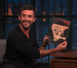 A man holds up a child’s drawing of a ghost with googly eyes, smiling while seated on a talk show set