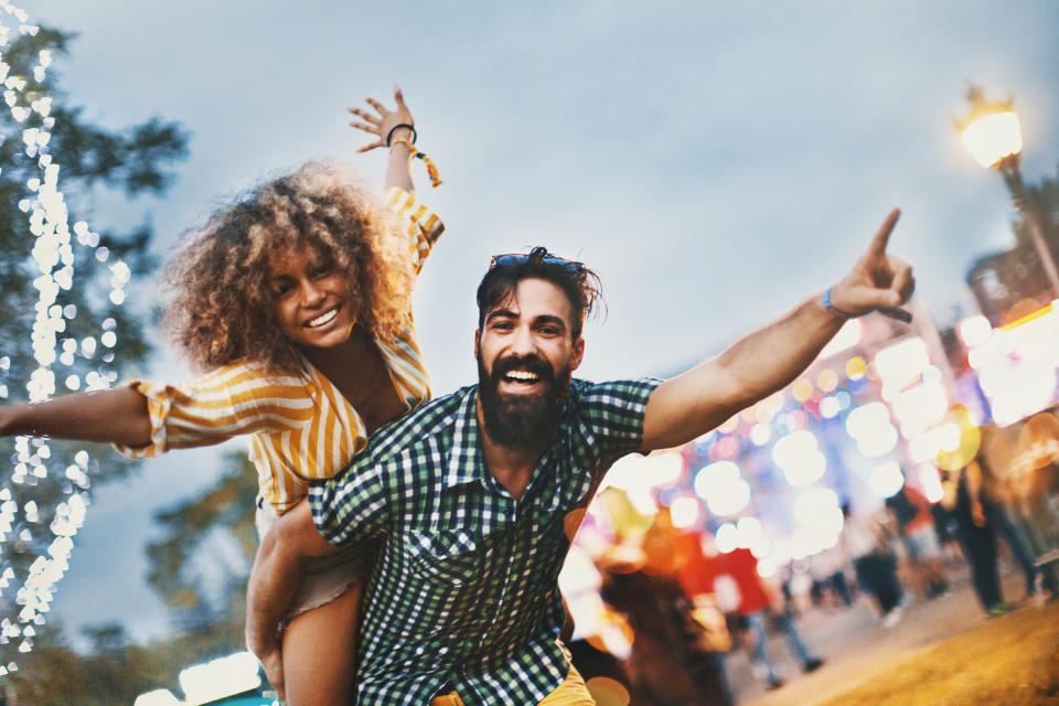 A woman with curly hair in a striped dress playfully rides on a man's back. Both are smiling with arms outstretched, surrounded by festival lights at night