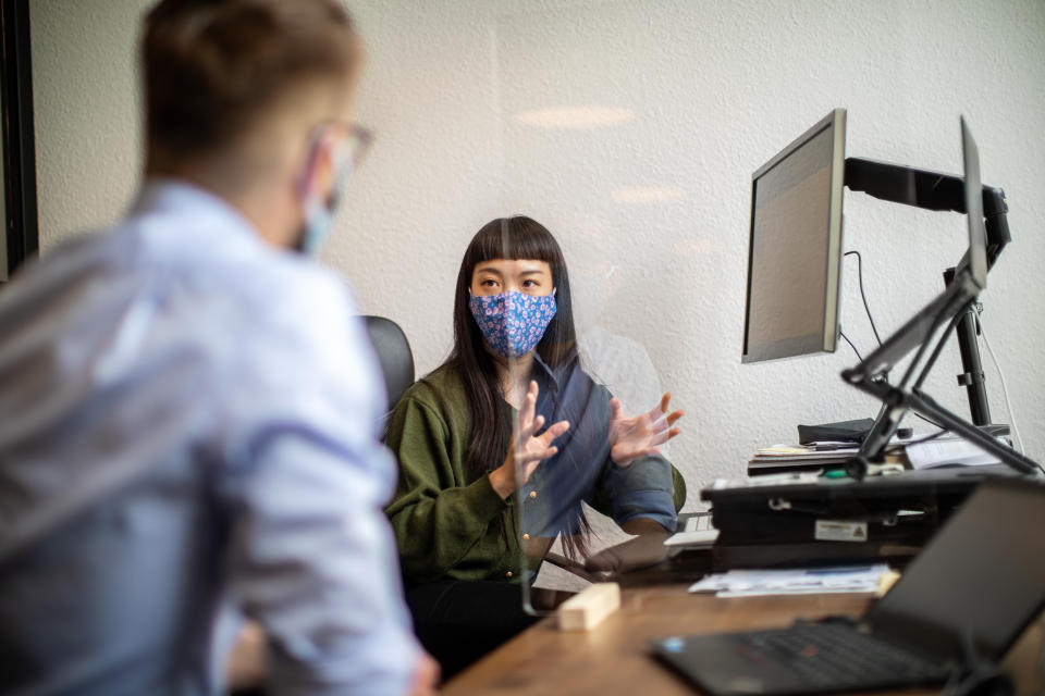 Businesswoman discussing work through glass partition with colleague at office desk. Business people back to work sitting at desk with protection guard between them post covid-19 pandemic.