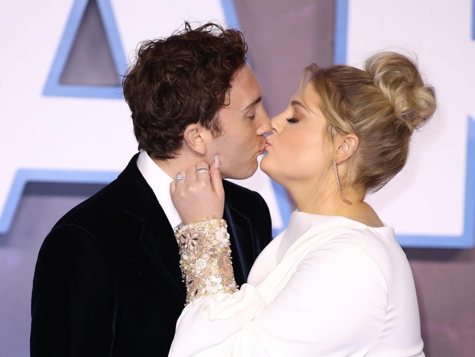 daryl sabara and meghan trainor kissing on the red carpet. sabara is wearing a black suit, while trainor is in a white dress with poofy sleeves and jewel encrusted cuffs. she has her hand held up to his face