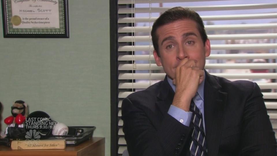 Steve Carell, dressed in a suit and tie, sits at a desk with a thoughtful expression, propping his chin on his hand. Office supplies are visible in the background