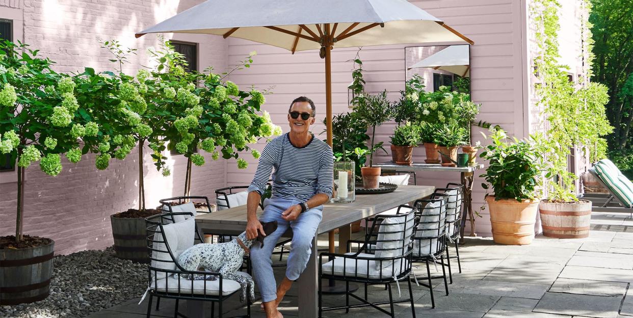 on a slate patio with flowering shrubs is a man sitting on a teak table with eight metal chairs with cushions, a dog seated on one, and a sun umbrella next to a brick and clapboard house painted light pink