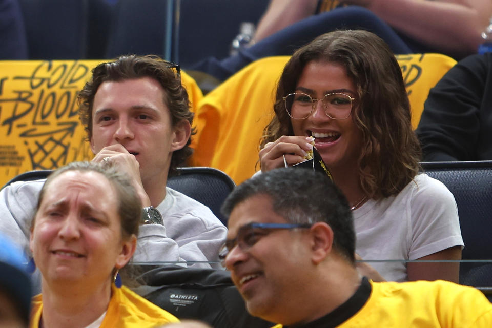 Tom Holland and Zendaya are seated together at a sports event, casually dressed, smiling and enjoying the game. Other spectators are visible around them