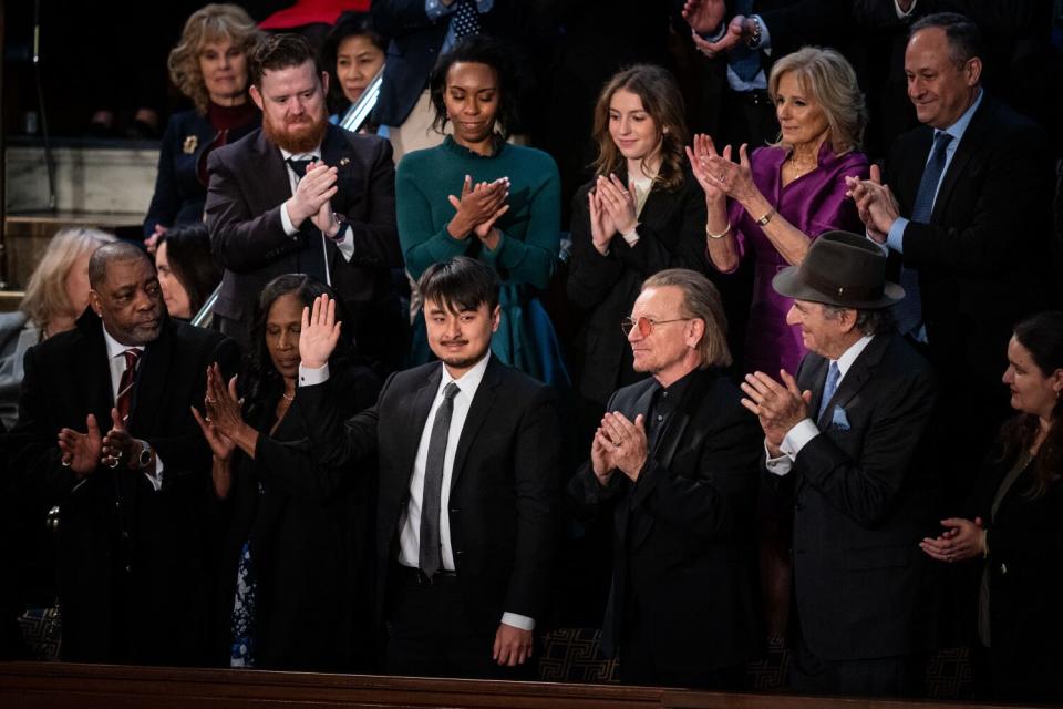 A group of well-dressed people applauds a young man in the middle as he raises his right hand in greeting.