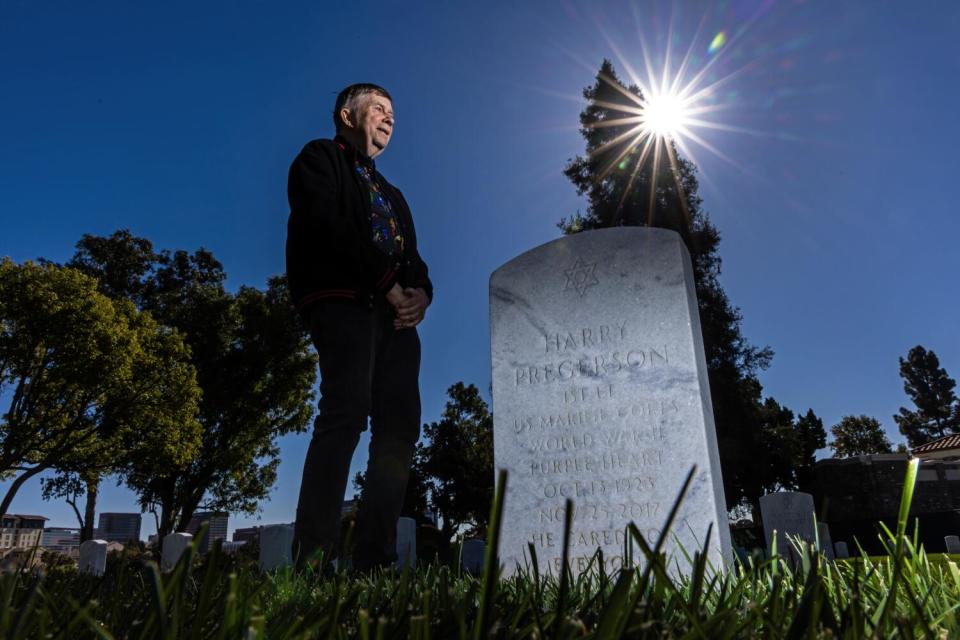 A man stands in near darkness by a headstone that bears a Star of David and reads "Harry Pregerson ... He cared for everyone"