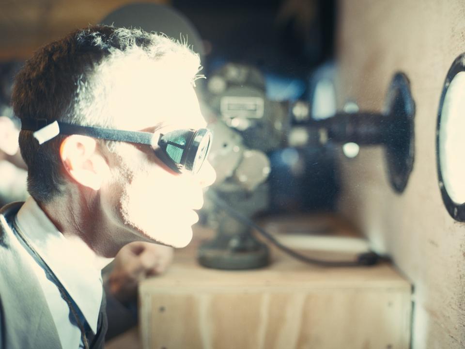 Cillian Murphy in a scene from "Oppenheimer" where Oppenheimer is seen wearing a set of googles and looking through a porthole while a bright light illuminates his face.