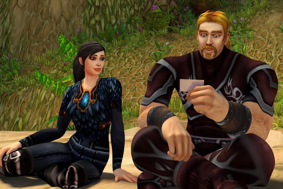 A video-game still shows two fantasy characters sitting down on a hillside, an attractive dark-haired woman and a burly blonde man in a goatee. She looks on as he reads a note in his hand.