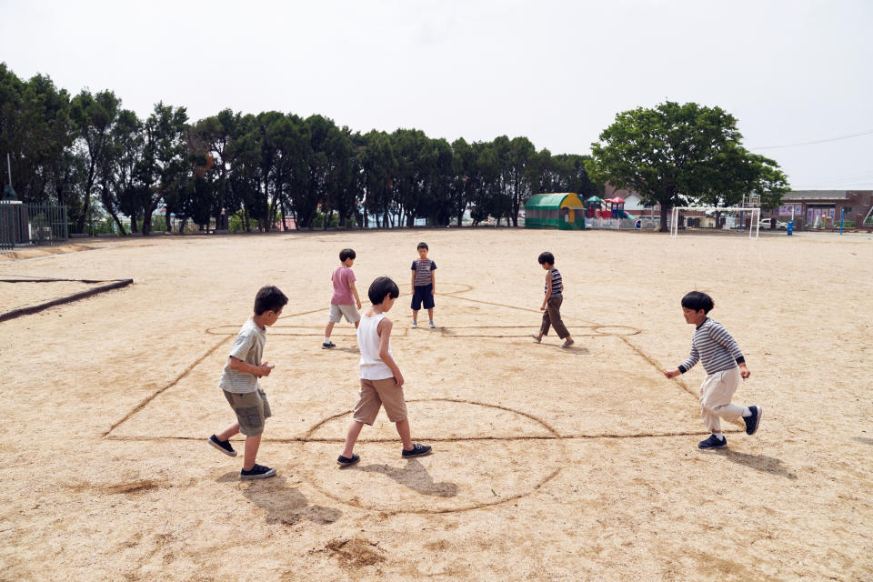 Children playing a game on a school playground drawn with lines, surrounded by trees and some distant buildings in the background