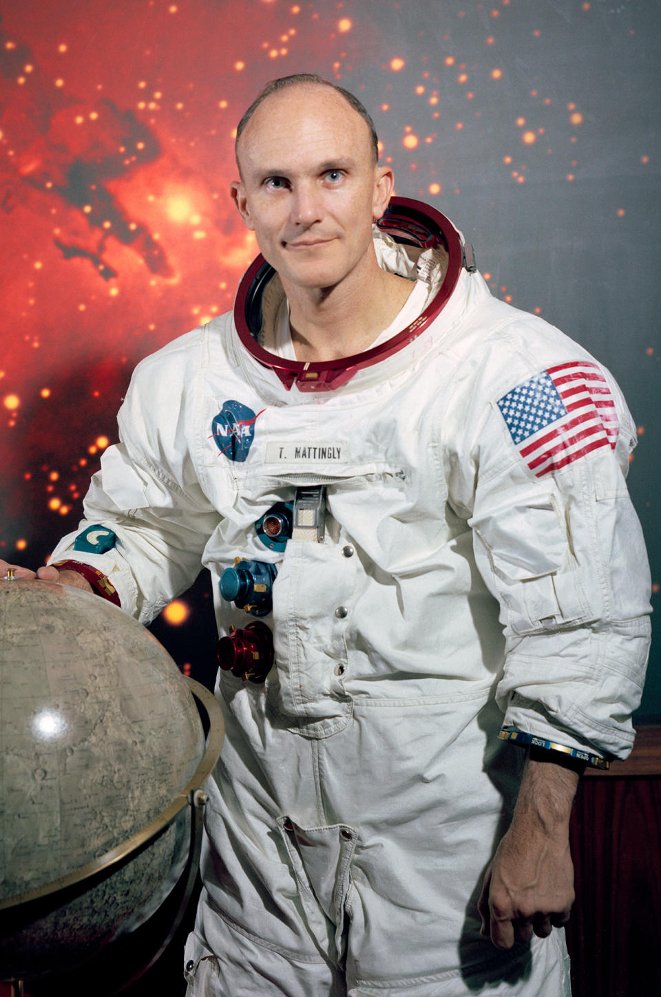 a man in a white spacesuit without a helmet stands near a model of the moon.