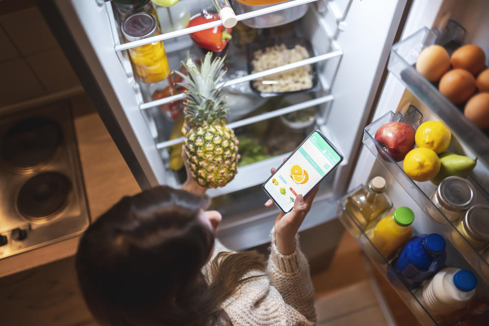 High angle view of a beautiful young woman standing next to an opened refrigerator door, holding a smart phone and ordering fresh fruit and vegetables online for home delivery