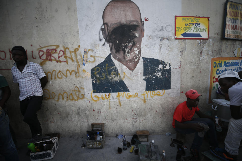 A man shines a client's shoes as another waits for business, in front of a wall painted with an image of President Jovenel Moise, his face obscured by black spray paint, in the Delmas neighborhood of Port-au-Prince, Haiti, Tuesday, Oct. 8, 2019. The country has entered its fourth week of anti-government protests that have paralyzed the economy and shuttered schools as demonstrators demand the resignation of the president. (AP Photo/Rebecca Blackwell)