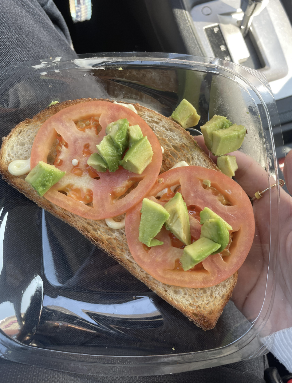 Close-up of a hand holding a clear container with a slice of bread topped with mayonnaise, tomato slices, and diced avocado