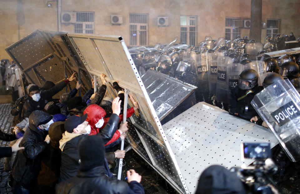 A group of protesters hold up a metal barricade against serried ranks of police with transparent shields and face guards.