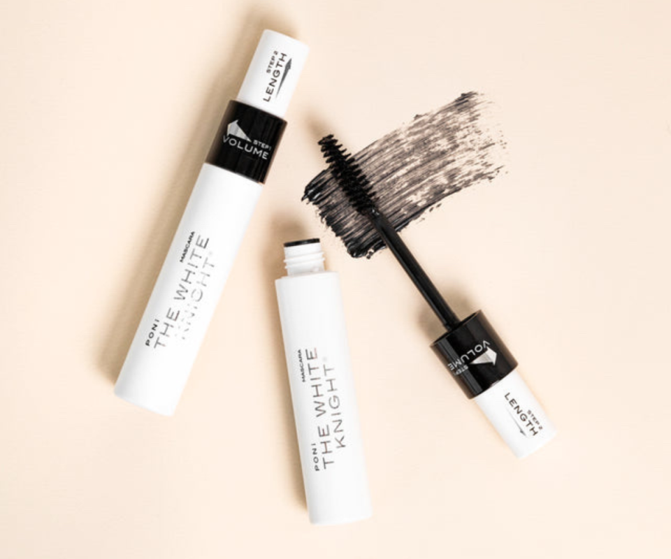 The White Knight mascara tube in white lying on a cream background with another tube on the right with the brush out and black formula smeared on the surface.