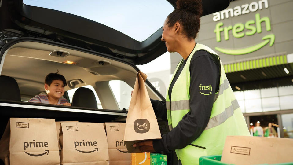 Ein Mitarbeiter von Amazon Fresh in einer neongrünen Weste lädt Prime Fresh-Lebensmittel in ein Fließheck, während auf dem Rücksitz ein Kind grinst.  Die Person, die die Lebensmittel einlädt, lächelt zurück. 