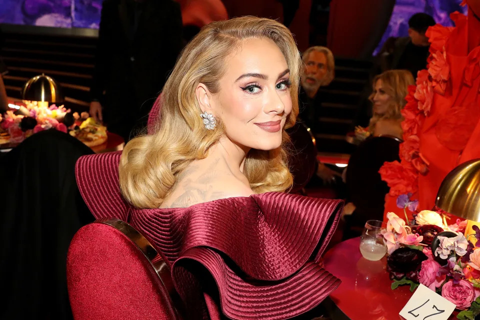 Adele sits at an event table, wearing a glamorous gown with extravagant ruffled shoulders, looking over her shoulder towards the camera