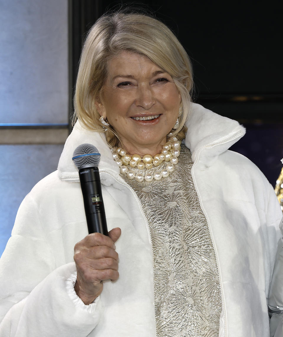 Martha Stewart in a white coat and pearl necklace, smiling and holding a microphone