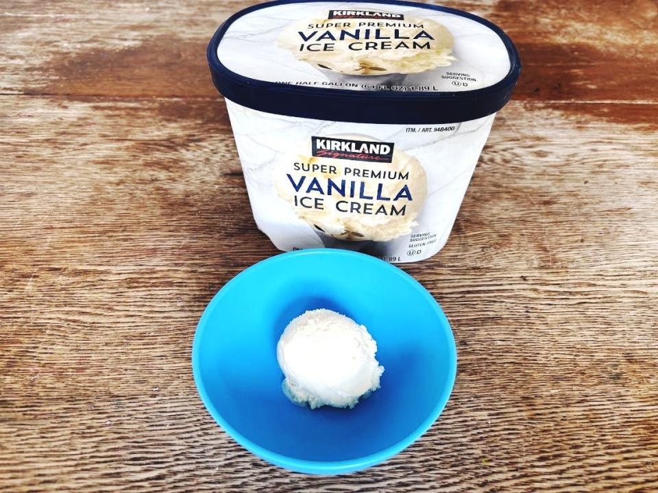 A gray carton of vanilla ice cream with a picture of a scoop of ice cream and Kirkland Signature logo on the front. A blue bowl holding a scoop of vanilla ice cream sits next to the carton