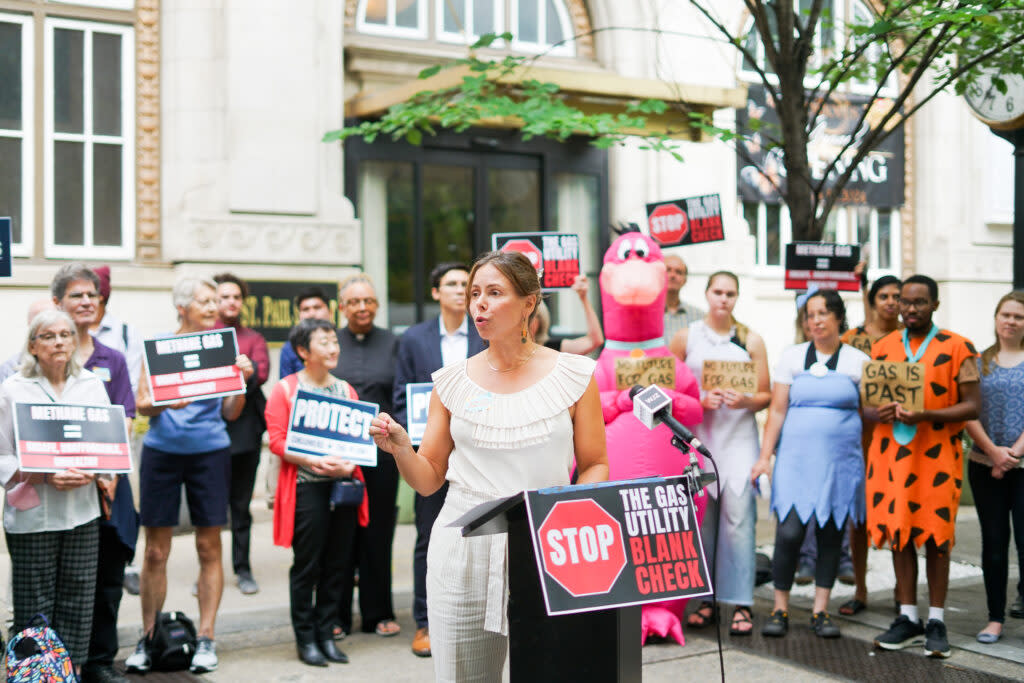 Advoacte Clara Vondrich speaking in front of a podium with advocates with signs behind her