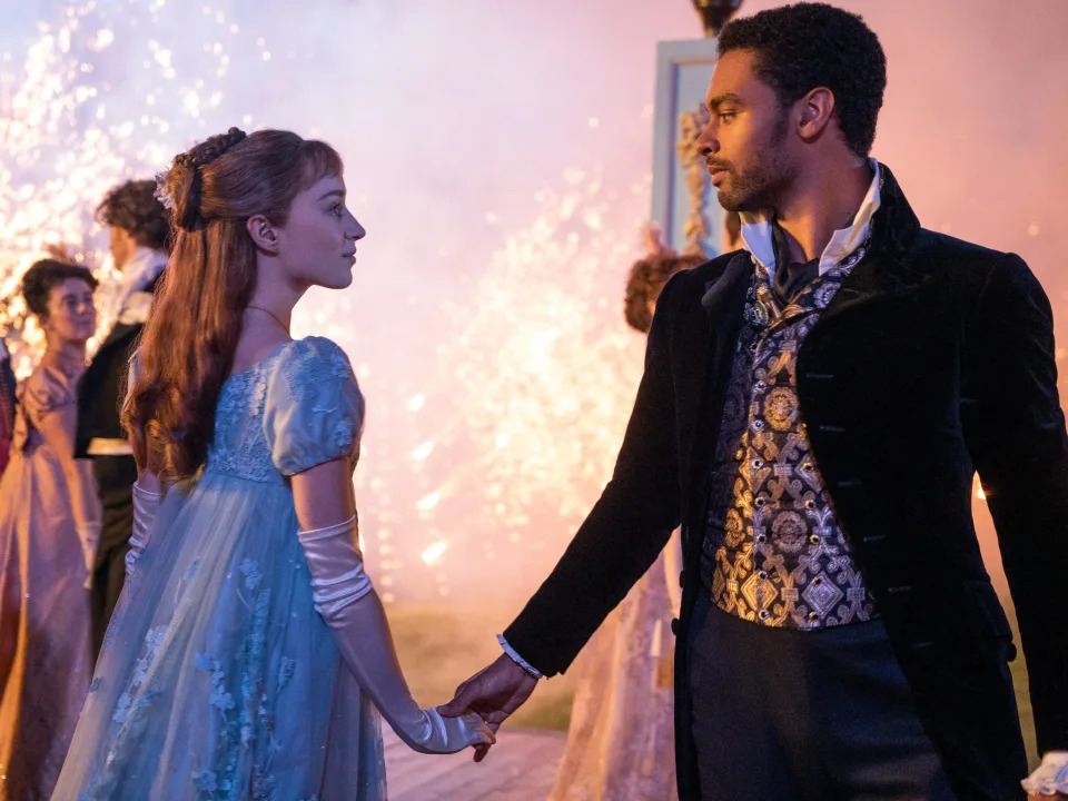 daphne and simon dancing in bridgerton. daphne is wearing a sheer blue gown, her hair worn half up, and simon is wearing a shining waistcoast and black overcoat. they are looking intently into each others eyes