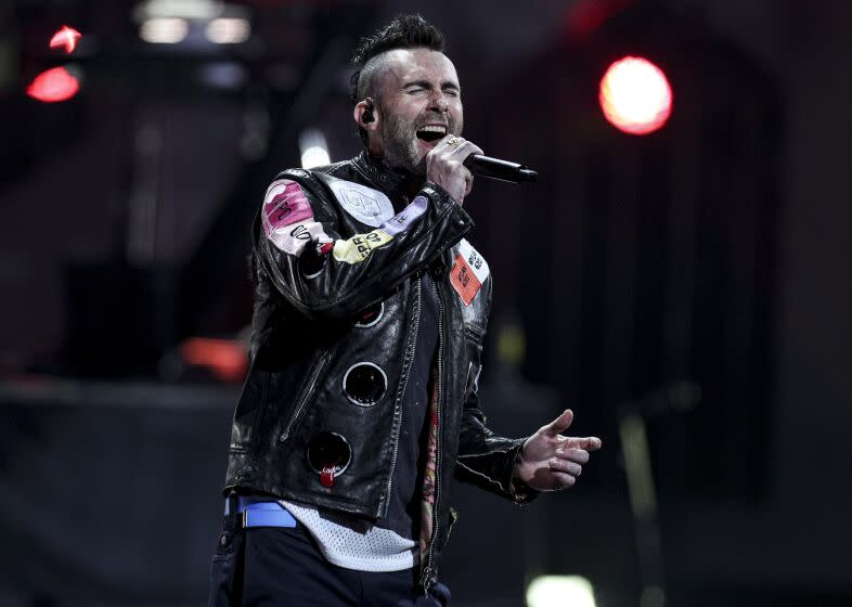 Adam Levine wearing a leather jacket with patches and singing into a microphone on stage