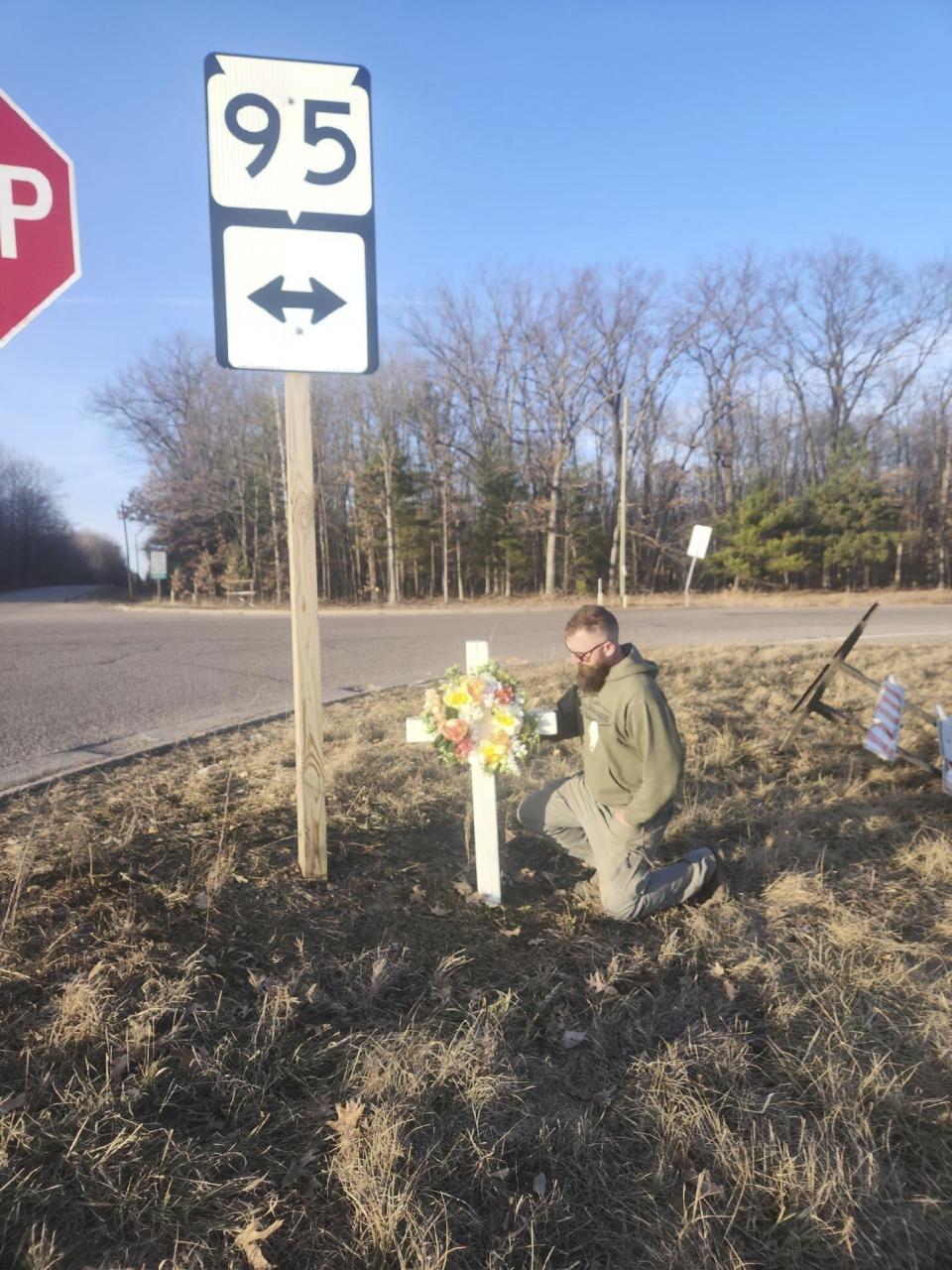 In this photo provided by Jennifer Jahn, her husband, Nathaniel Jahn, places a wood cross with a flower wreath at the site of a crash that killed nine people in Clark County, Wis., Sunday, March 10, 2024. Nathaniel Jahn witnessed the crash and then ran over to try to help on March 8 on his way to work. He ended up saving a 2-year-old boy. (Jennifer Jahn via AP)