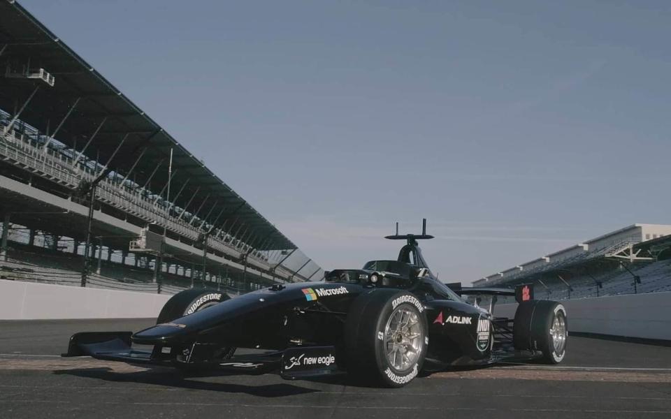 A sleek black Indy-standard car sits on a race track, with a package of cameras and other sensors installed where the driver would normally sit and a radar antenna mounted on top - Indy Autonomous Challenge