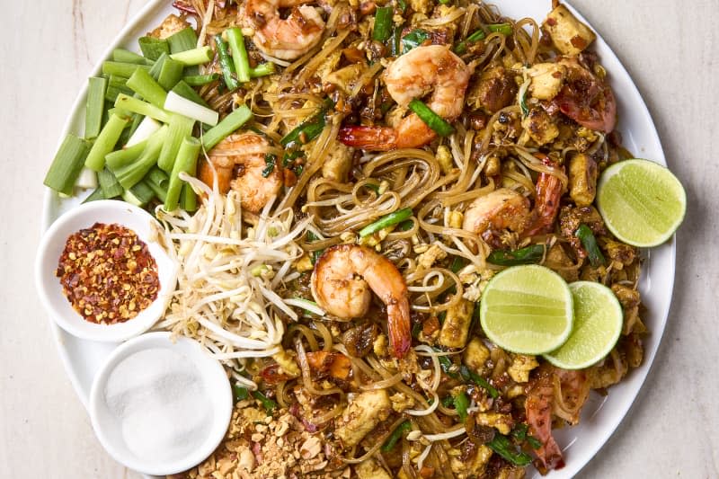 overhead shot of pad thai on a large white round plate, garnished with lime halves and green onion and a side of red pepper flakes and salt