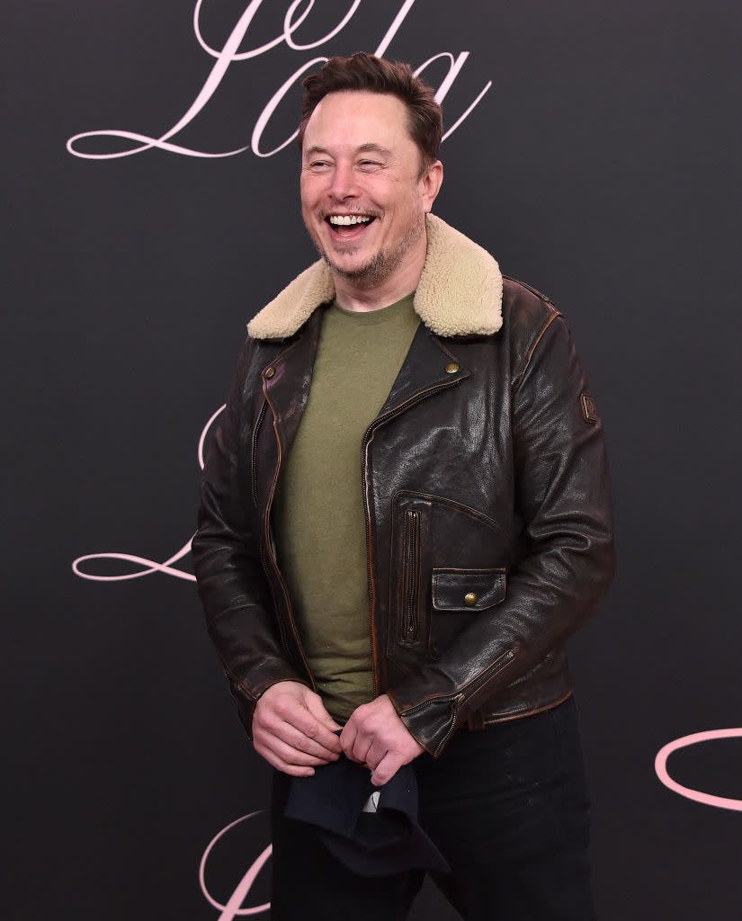 Elon Musk laughing in a leather jacket with a shearling collar and a green T-shirt