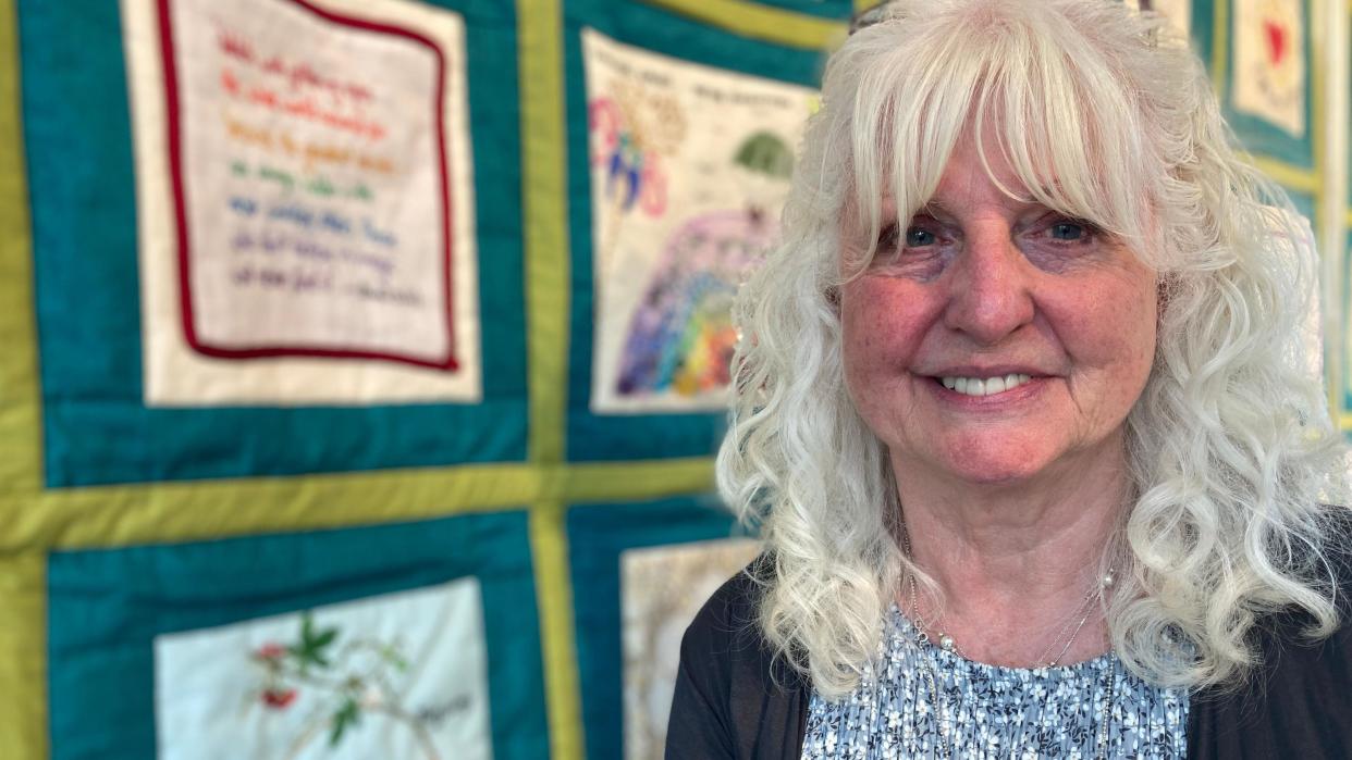 Beeb Deacon stands in front of the quilt with green and blue squares with individual panels within them