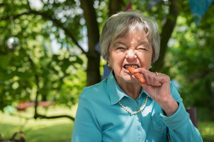 Elderly woman in a blue shirt and pearls biting a chicken wing with a joyful expression, outdoors
