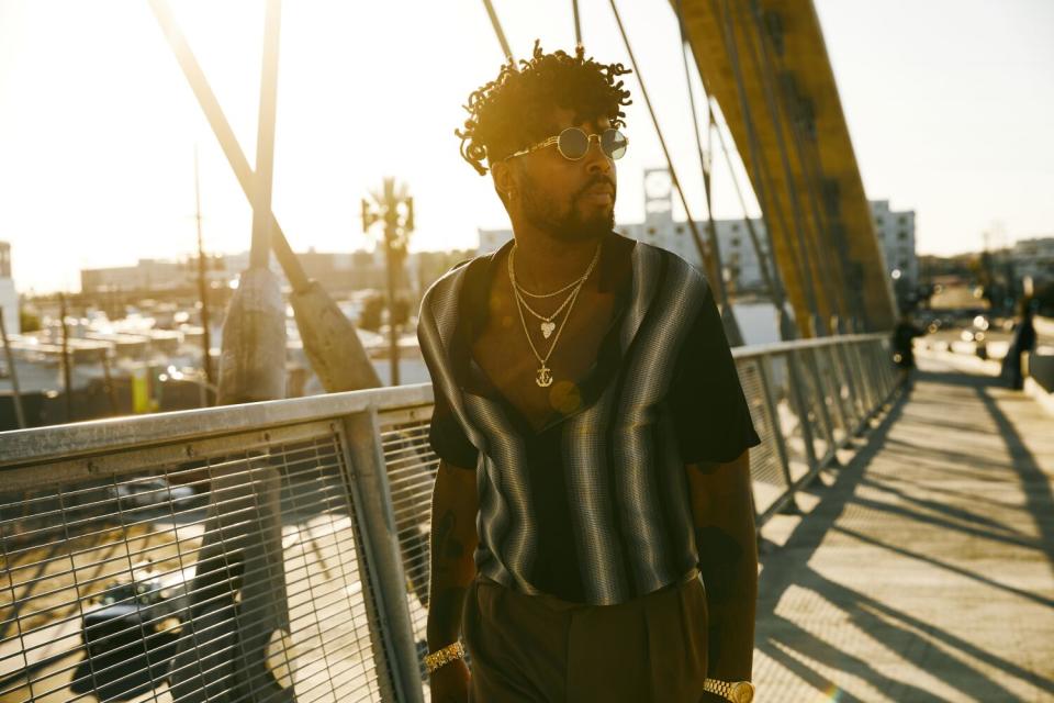 A man stands on a wooden walkway in a harbor, the sun behind him.