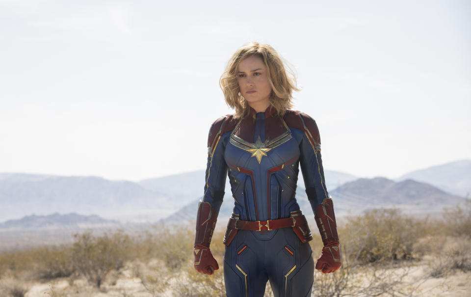 A blonde-haired woman in a red and white superhero costume standing in a dessert.