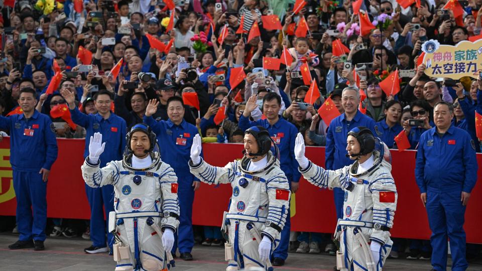 Three astronauts in white spacesuits wave red flags to a smiling crowd