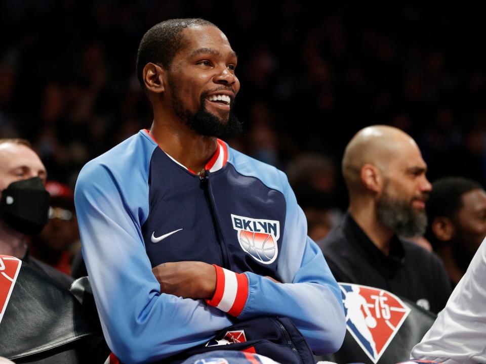 Kevin Durant crosses his arms and smiles while sitting on the bench in warmups.