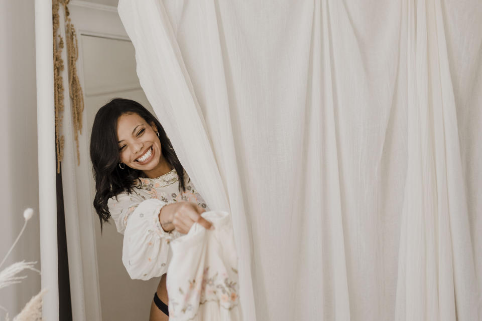 A woman, wearing a floral blouse, smiles while holding a white curtain in a room with neutral decor