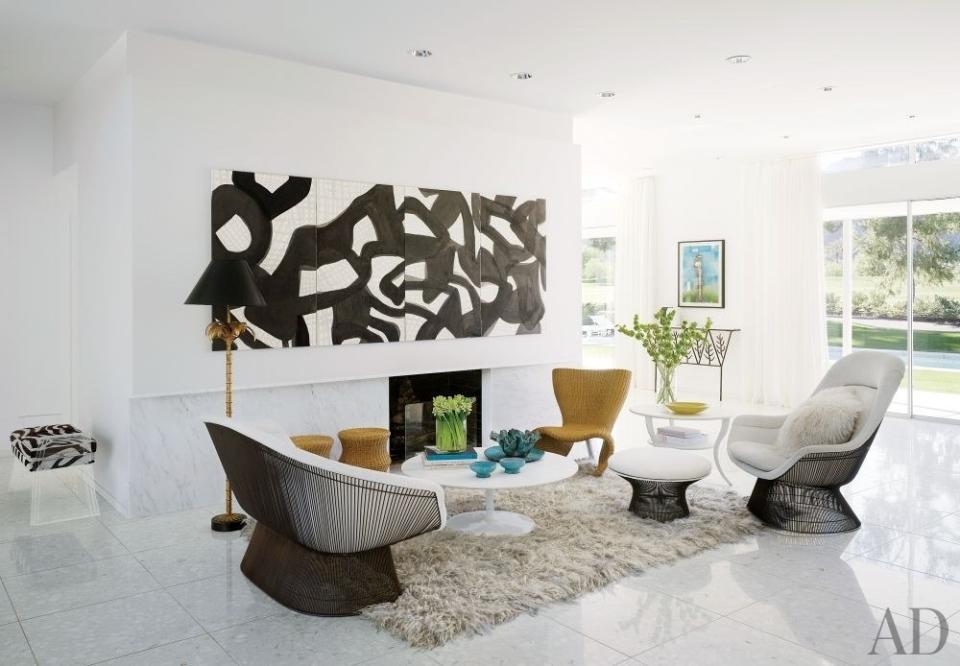 A living room in the California desert features a Warren Platner settee, lounge chair, and ottoman, all upholstered in a fabric from Cowtan & Tout, and a wicker chair by Marc Newson; the panels over the fireplace, clad in a hand-painted Porter Teleo wallpaper, conceal a television. Between the glass doors is an Eric Schmitt console, surmounted by a Jim Dine lithograph. The rug is by Madeline Weinrib, and the walls are painted in a Benjamin Moore white.